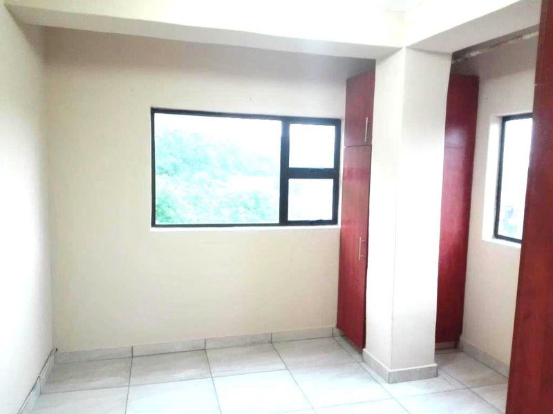A bright, empty bedroom featuring tiled flooring, built-in wooden cupboards, and large windows providing natural light.