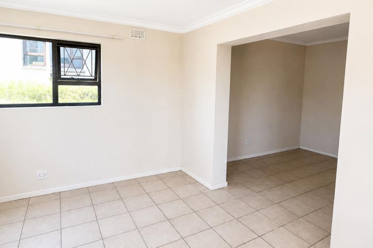 An empty, neutrally painted living area featuring tiled flooring, large windows with security bars, and a seamless transition into an adjacent room.
