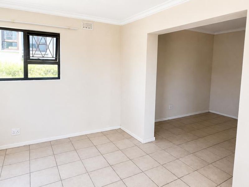 An empty, neutrally painted living area featuring tiled flooring, large windows with security bars, and a seamless transition into an adjacent room.