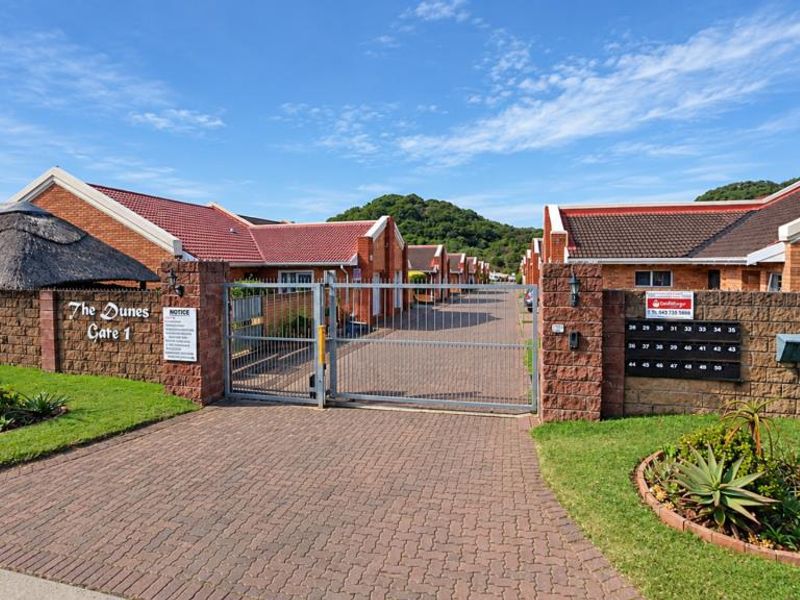 Front entrance view of The Dunes complex featuring a secure gated access point, brick boundary walls, and paved driveway leading to townhouse units.