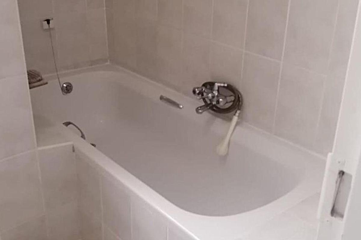 A functional bathroom featuring a built-in bathtub with wall-mounted mixer tap and handheld shower attachment, finished with neutral ceramic wall tiling.