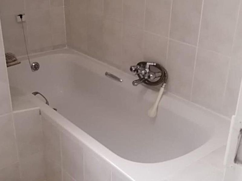 A functional bathroom featuring a built-in bathtub with wall-mounted mixer tap and handheld shower attachment, finished with neutral ceramic wall tiling.
