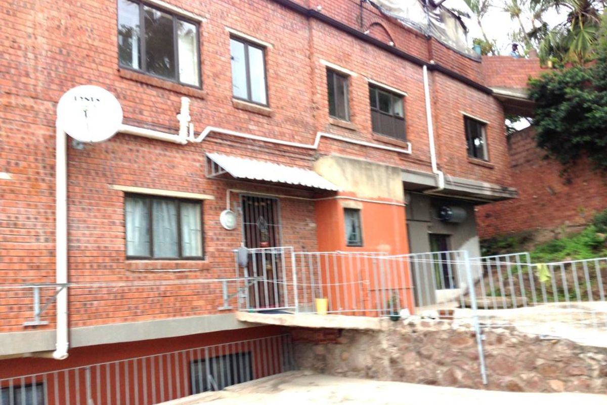 Exterior view of a multi-level brick apartment building featuring metal security gates, external plumbing, and a concrete patio area.