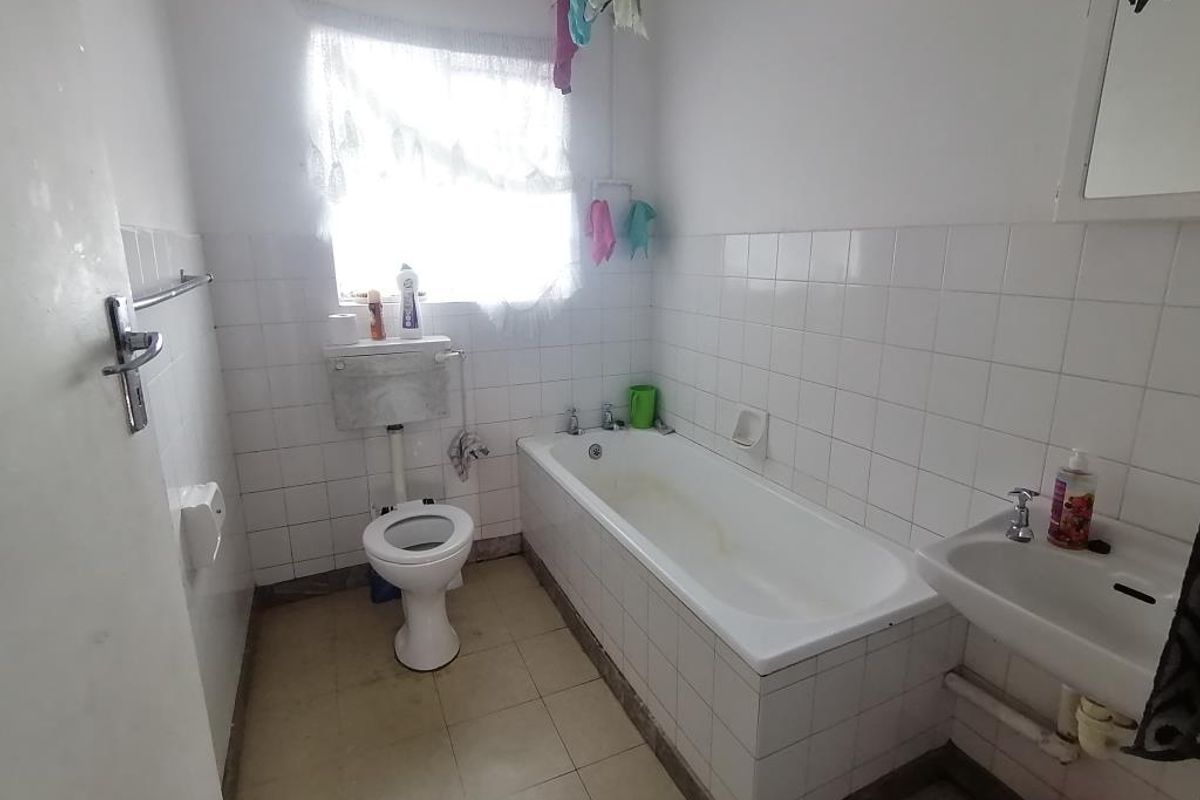 A functional bathroom featuring a bathtub, toilet, and washbasin with white wall tiling and tiled flooring, offering potential for modernization.