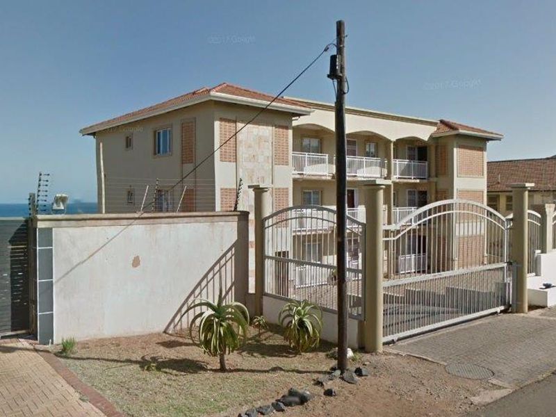 Exterior view of a multi-story apartment complex featuring a gated entrance, perimeter security fencing, and balconies overlooking the coastal area.
