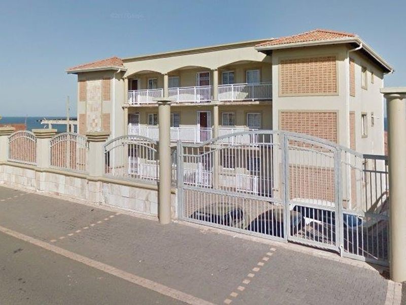 Exterior view of a multi-story apartment building featuring a secure gated entrance, perimeter fencing, and balconies overlooking the coastal area.