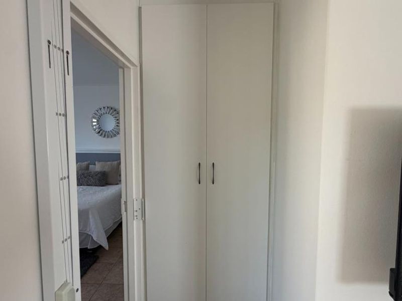 A clean, empty bedroom featuring neutral tiled flooring, built-in white cupboards, and an aluminium-framed window with security bars.