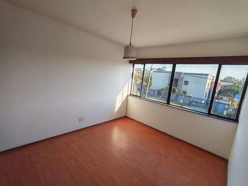 A bright, empty bedroom featuring laminate flooring, a large window with natural light, and a simple pendant light fixture.