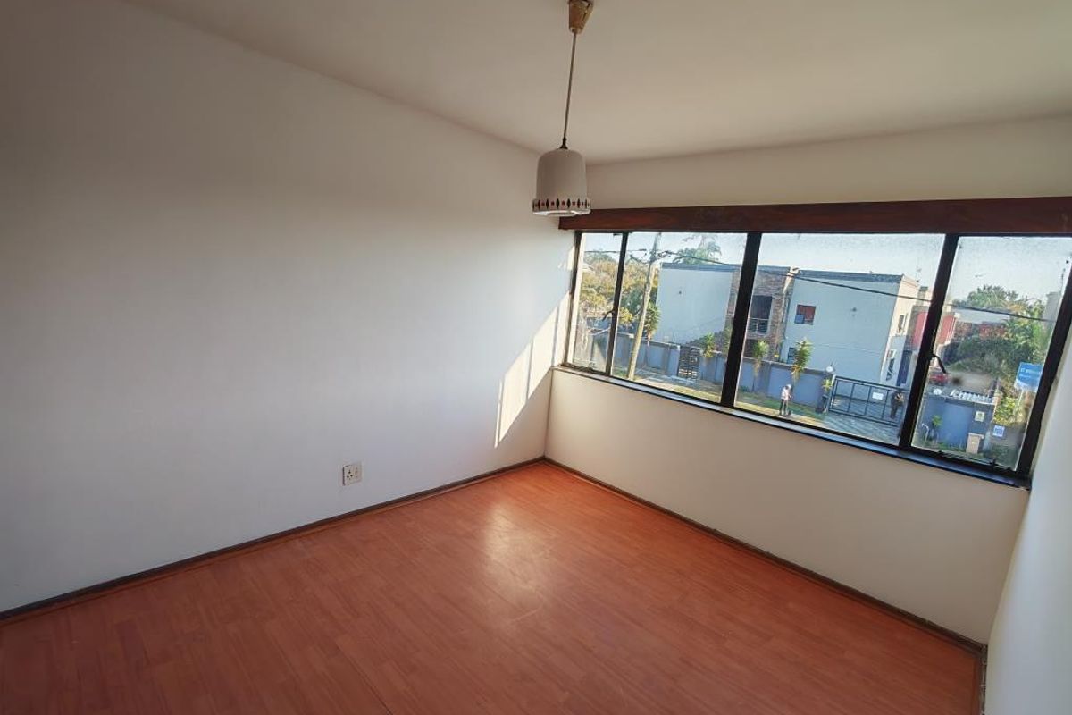 A bright, empty bedroom featuring laminate flooring, a large window with natural light, and a simple pendant light fixture.