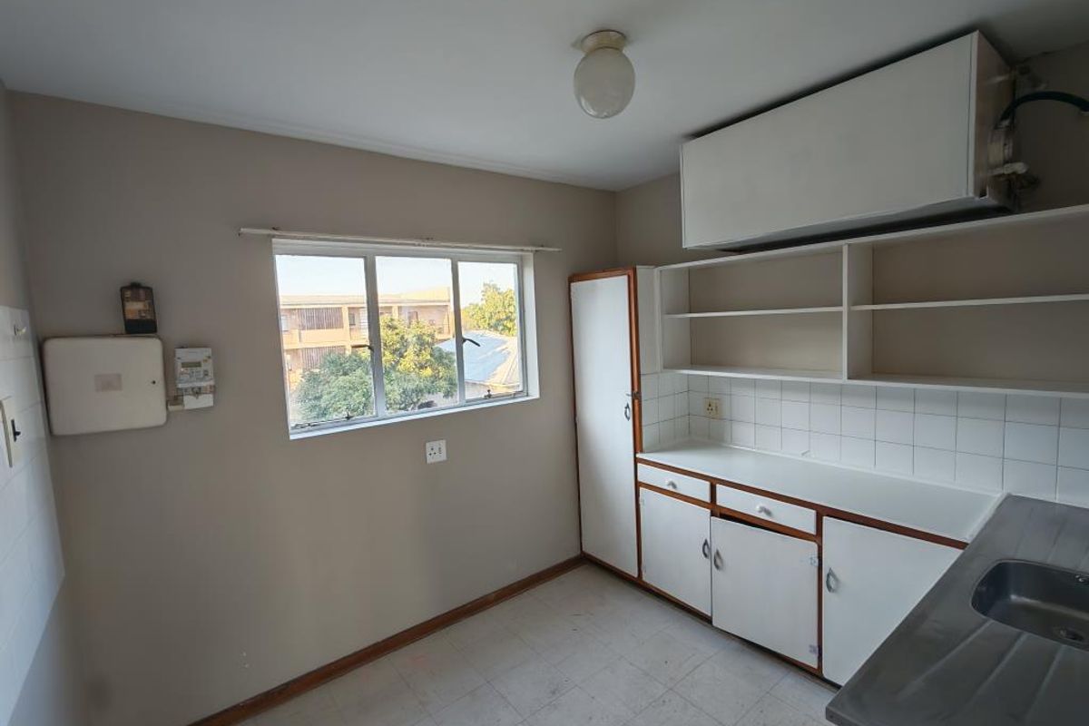 A functional, empty kitchen featuring built-in cabinetry, open shelving, a stainless steel sink, and a tiled backsplash, offering potential for modernization.