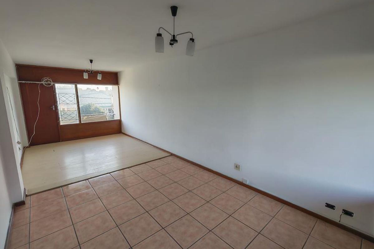 An empty, open-plan living area featuring a combination of tiled and laminate flooring, neutral walls, and access to a balcony through a security gate.