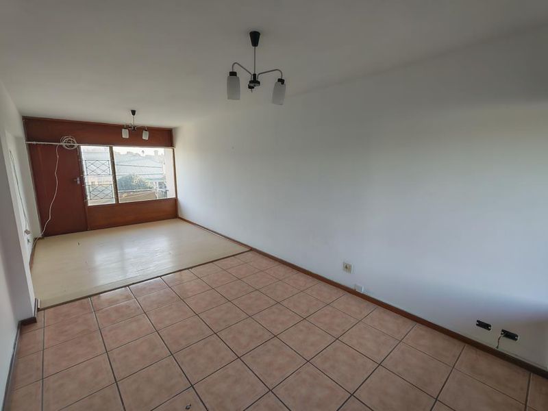 An empty, open-plan living area featuring a combination of tiled and laminate flooring, neutral walls, and access to a balcony through a security gate.