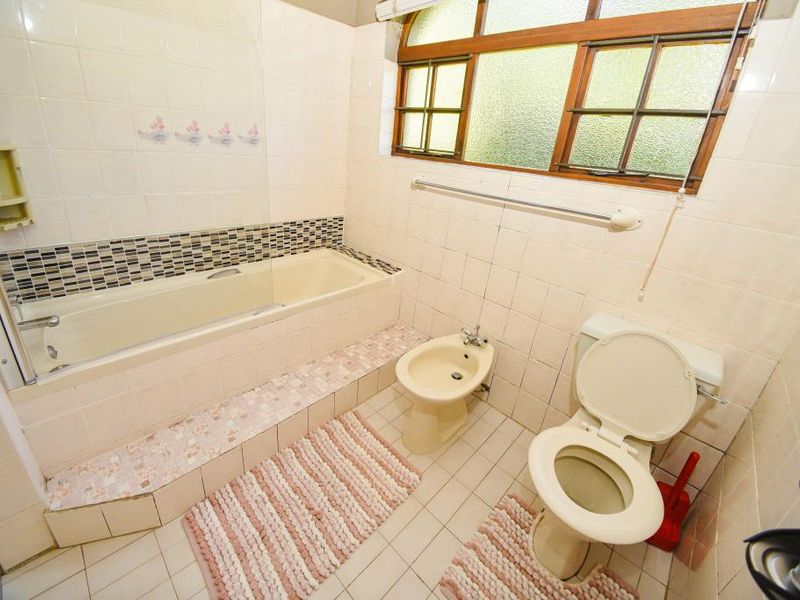 A dated bathroom featuring a built-in bathtub with shower screen, toilet, and bidet, finished with original wall and floor tiling.