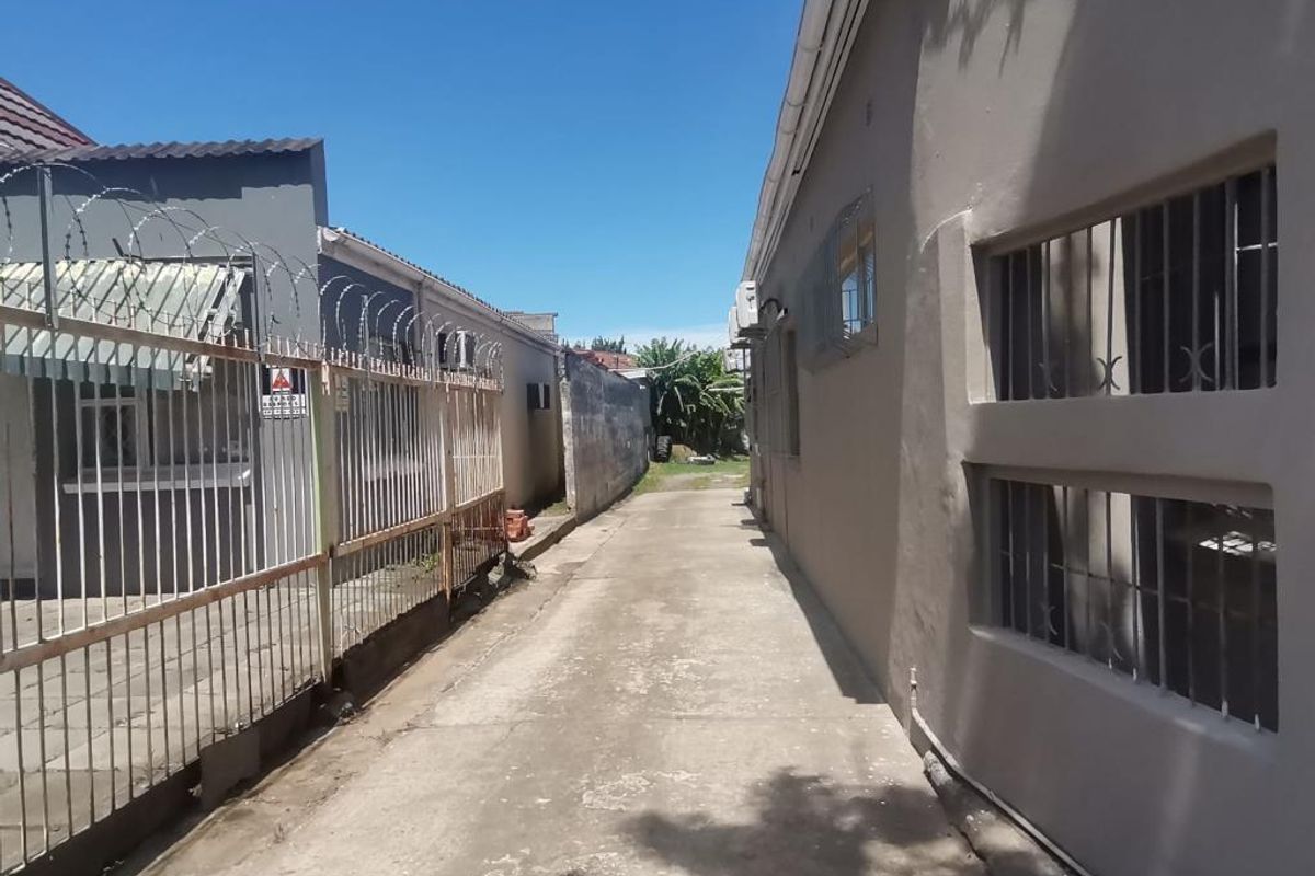 A narrow concrete side alleyway between two commercial buildings, featuring security fencing and metal window grilles.