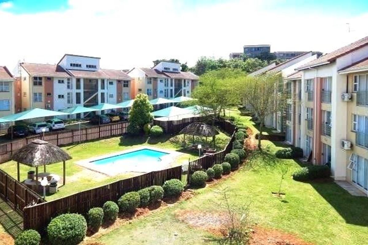 Elevated view of the apartment complex grounds, featuring a communal swimming pool, thatched gazebo, landscaped lawns, and covered parking bays.