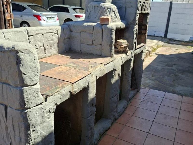 Outdoor built-in stone braai and barbecue area featuring a chimney and integrated storage compartments, set on a tiled patio surface.