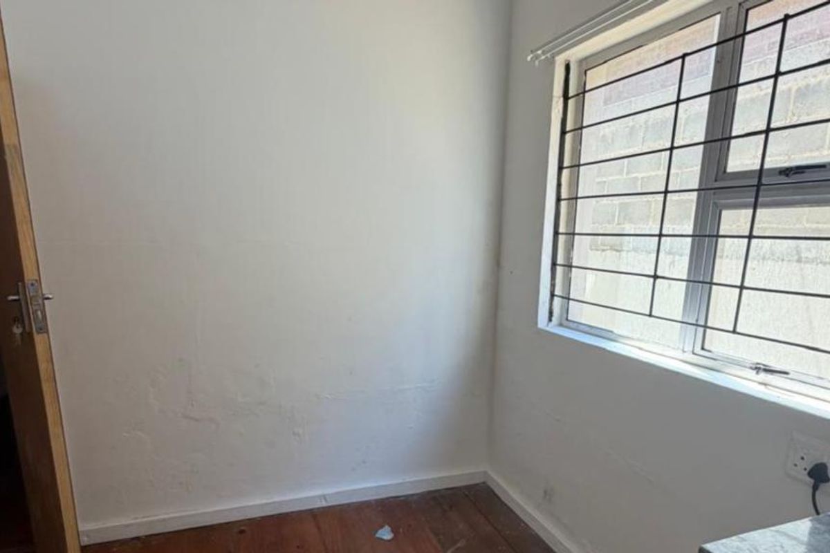 An empty bedroom featuring timber flooring, a window with security bars, and visible wall surface wear requiring maintenance.
