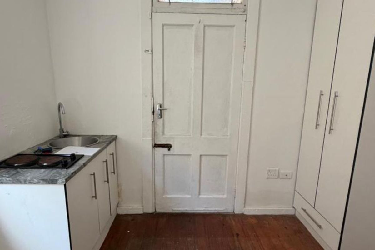 A compact kitchenette area featuring a small sink, electric hob, built-in cabinetry, and a tall wardrobe, set against a backdrop of timber flooring.