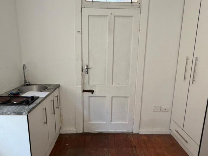 A compact kitchenette area featuring a small sink, electric hob, built-in cabinetry, and a tall wardrobe, set against a backdrop of timber flooring.