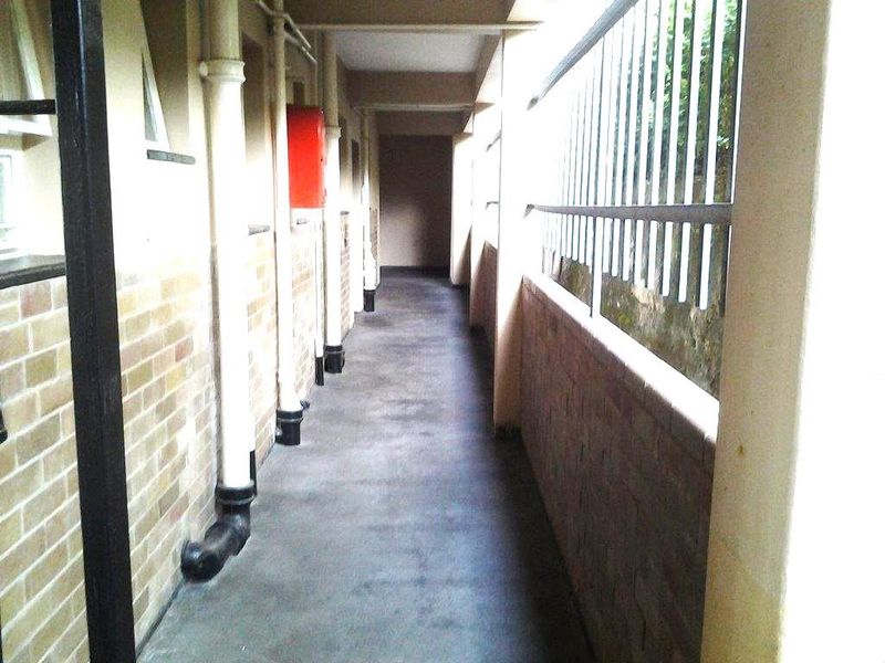 A utilitarian apartment building hallway featuring exposed plumbing, brick walls, and metal security railings.