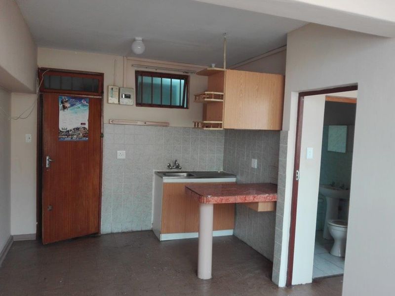 A compact kitchen area featuring basic cabinetry, a stainless steel sink, and a breakfast counter, with an adjacent doorway leading to a bathroom.