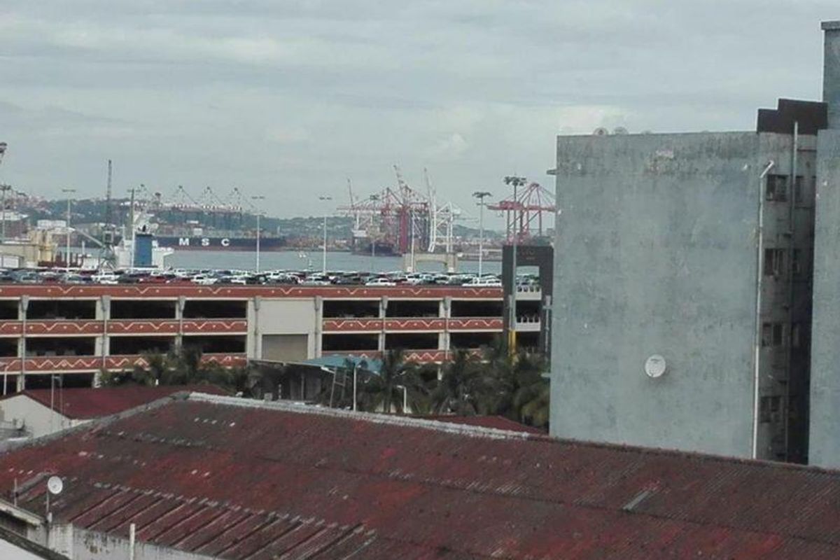 An elevated view overlooking a commercial harbor area with industrial cranes, shipping vessels, and a multi-level parking structure.