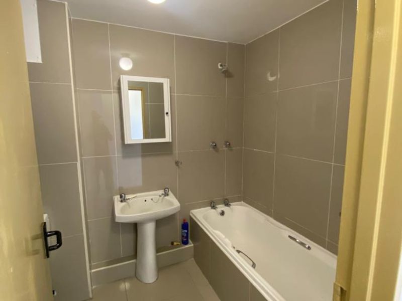 A functional bathroom featuring floor-to-ceiling neutral tiling, a pedestal basin, and a standard bathtub with wall-mounted taps.