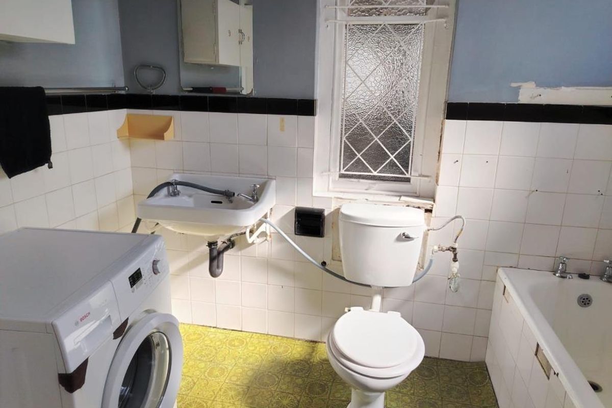 A dated bathroom featuring a bathtub, wall-mounted basin, and toilet, with visible wear to wall tiles and flooring, requiring renovation.