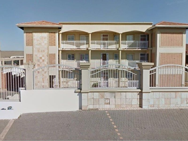 Front elevation of a multi-story apartment building featuring a paved driveway, secure perimeter fencing, and private balconies for each unit.