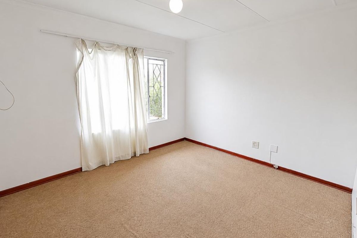 A bright, empty bedroom featuring neutral walls, carpeted flooring, and a window with security bars.