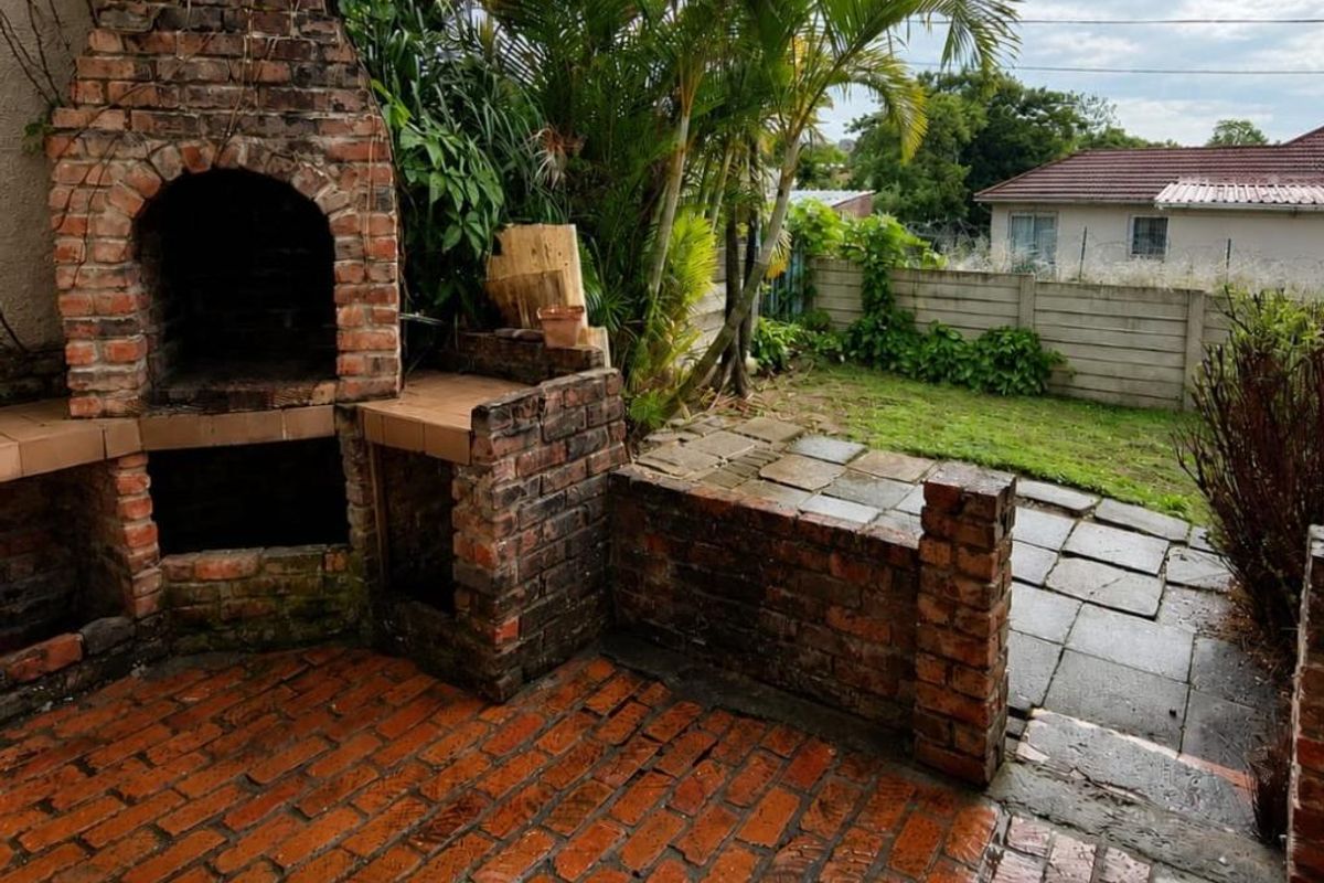 Outdoor patio area featuring a built-in brick braai and fireplace, leading onto a small garden space with paved walkways.