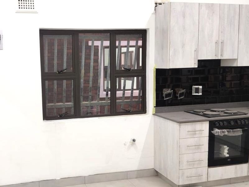 A compact kitchen featuring light wood-grain cabinetry, a black subway tile backsplash, built-in oven and hob, and security bars on the window.