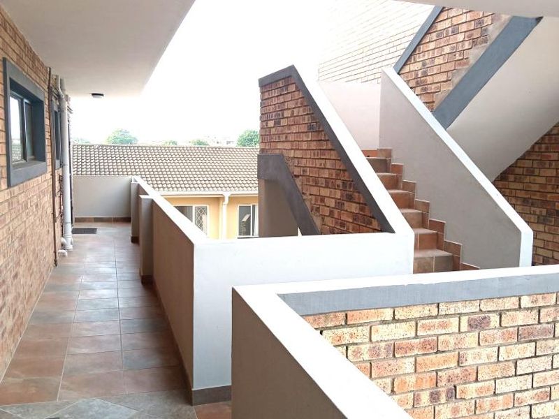 An open-air apartment hallway featuring exposed brick walls, tiled flooring, and a staircase leading to other levels.