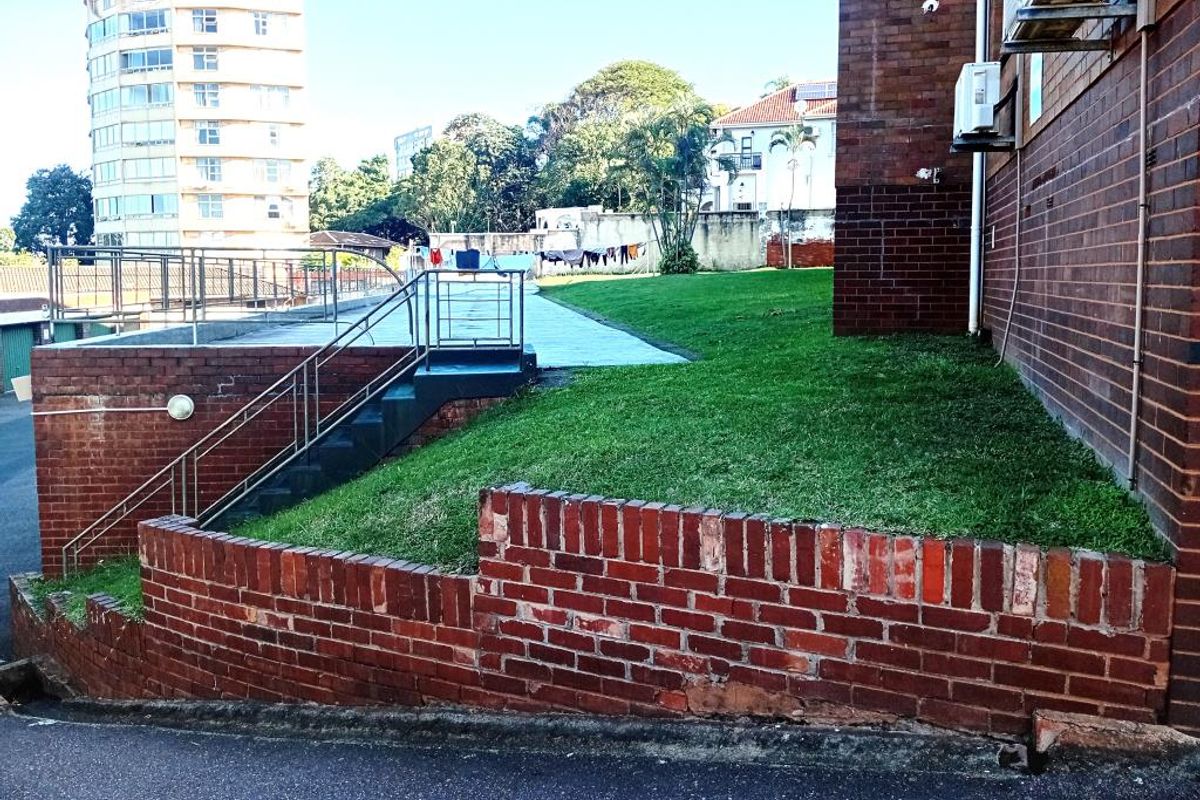 Exterior view of the apartment building featuring a brick facade, a grassed embankment, and a paved walkway with metal railings.