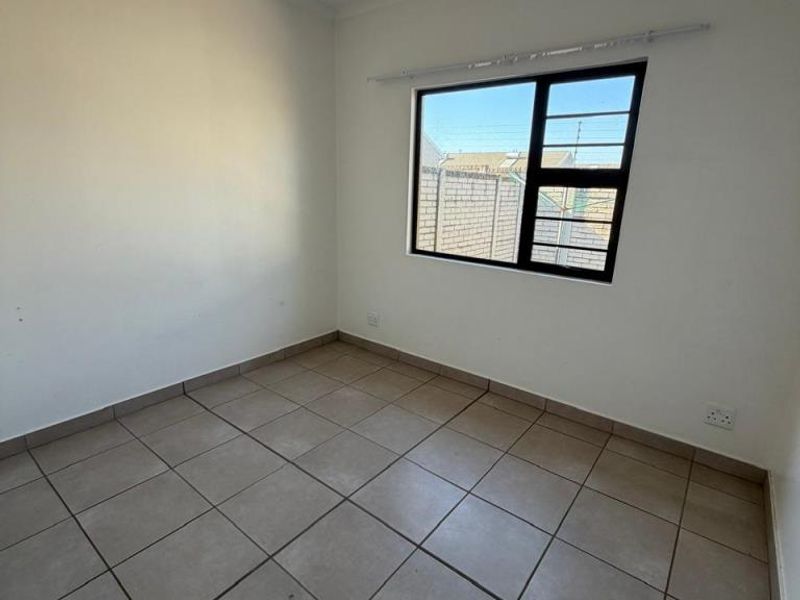 A bright, empty bedroom featuring neutral ceramic floor tiles, white walls, and a black-framed window with security bars.