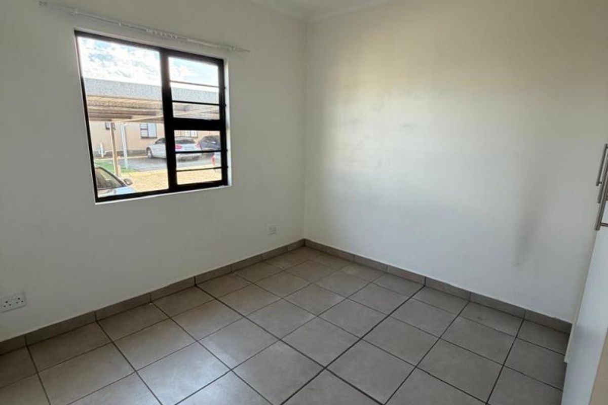 A bright, empty bedroom featuring neutral floor tiles, a large window with security bars, and built-in cupboard space.