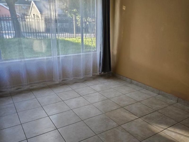An empty living room featuring neutral tiled flooring, a ceiling fan, and a large window with security bars overlooking the exterior.