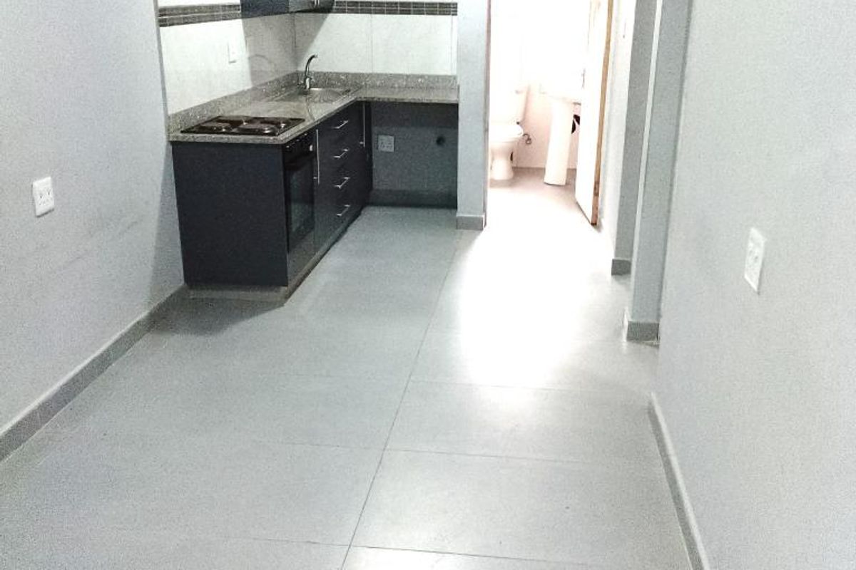 A compact kitchen featuring dark built-in cabinetry, granite countertops, a built-in oven and hob, and tiled flooring, leading to an adjacent bathroom.