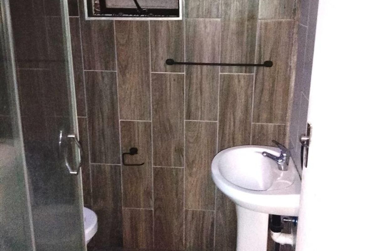 A compact bathroom featuring a pedestal sink, shower enclosure, and wood-look wall tiling with a louvered window for ventilation.
