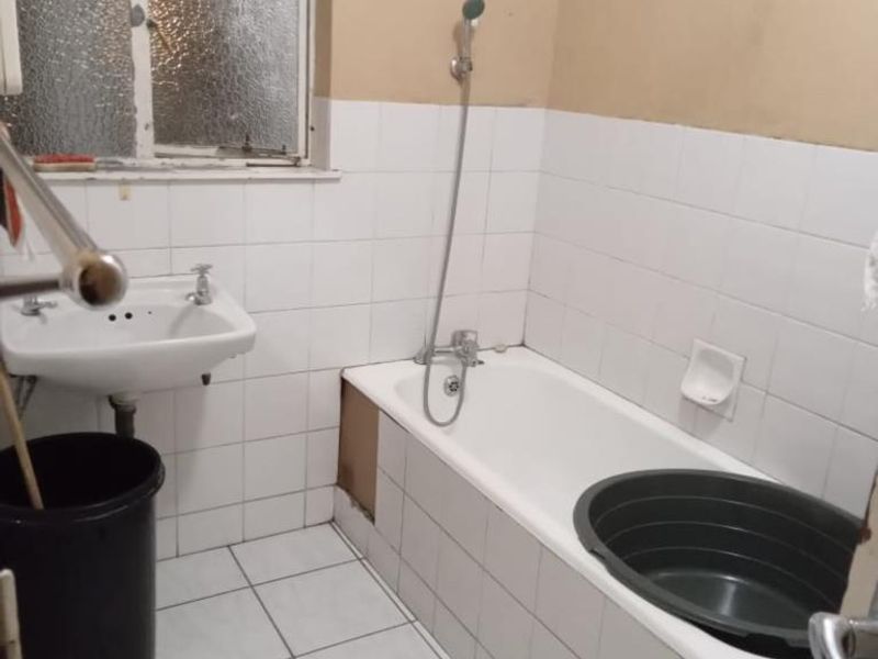 A dated bathroom featuring a bathtub with a handheld shower, a wall-mounted basin, and tiled finishes, currently requiring cosmetic updates and repairs.