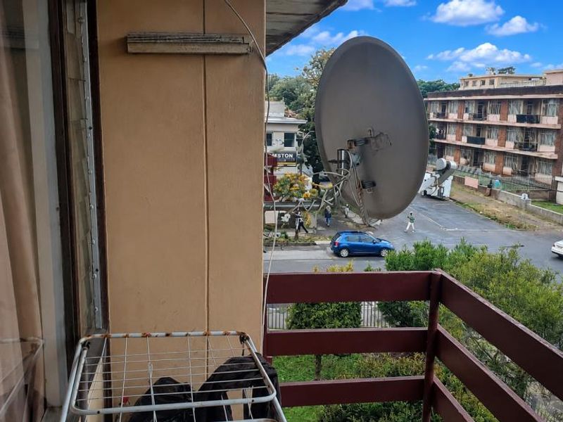 A balcony area featuring a timber balustrade and a wall-mounted satellite dish, overlooking a street and neighboring apartment buildings.