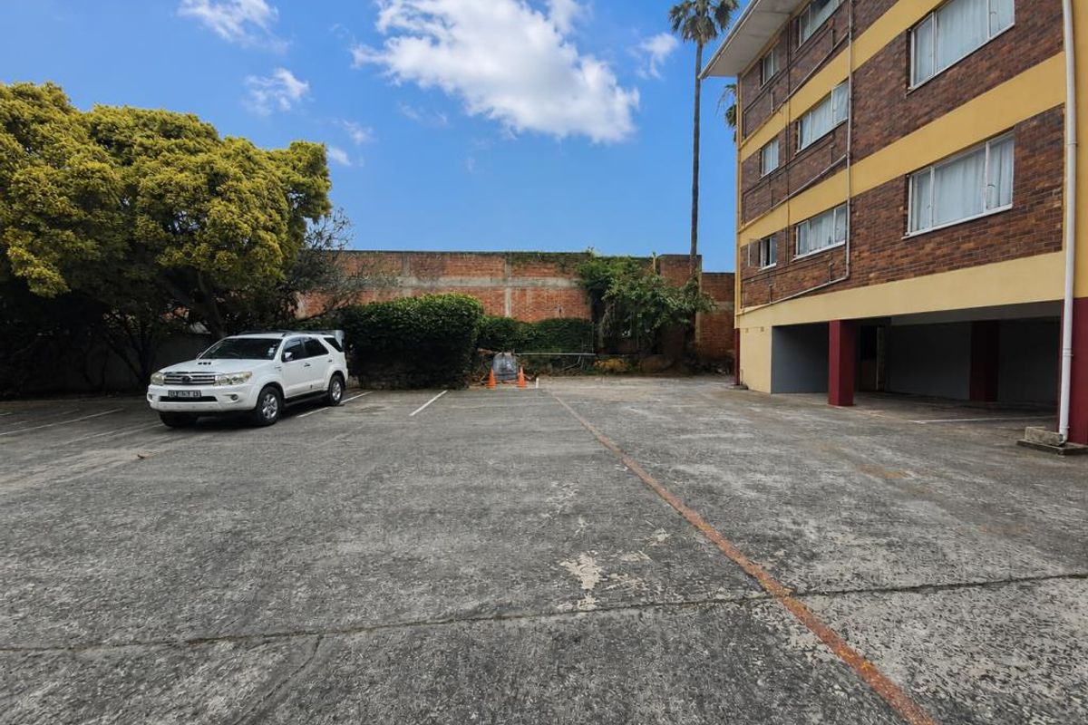 Rear exterior view of the apartment building featuring a concrete parking area with covered bays and a boundary wall.
