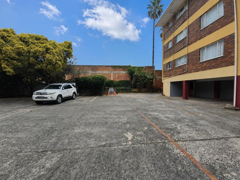 Rear exterior view of the apartment building featuring a concrete parking area with covered bays and a boundary wall.