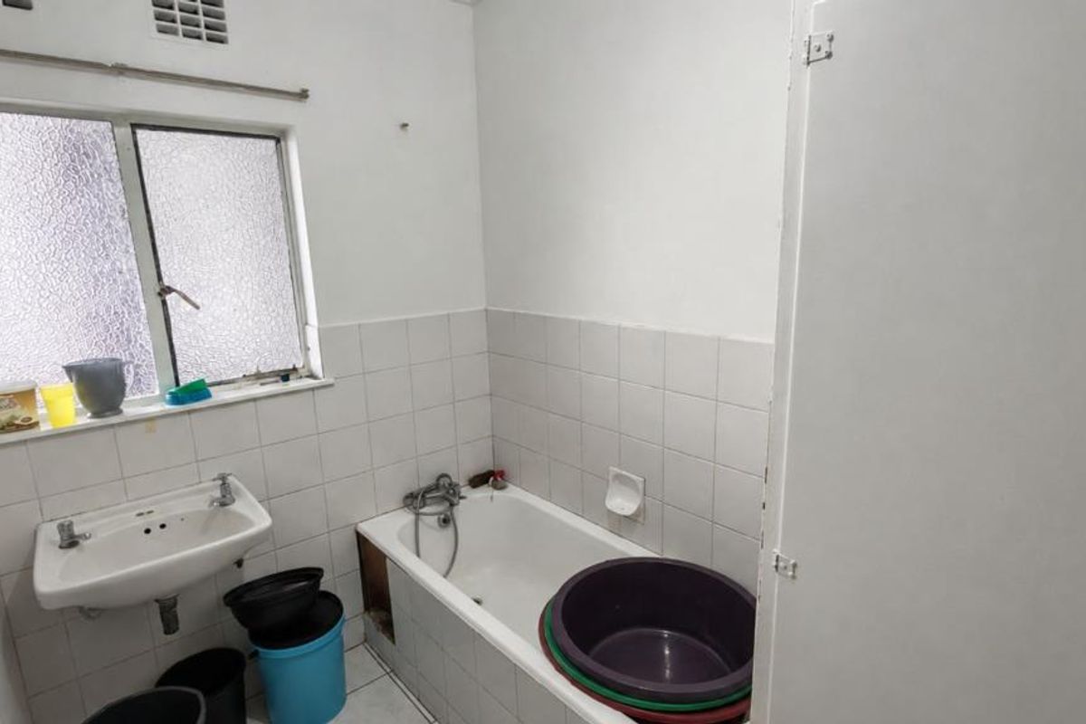 A functional bathroom featuring a bathtub, wall-mounted basin, and tiled finishes. The space requires cosmetic updates and repairs to the bathtub panel.