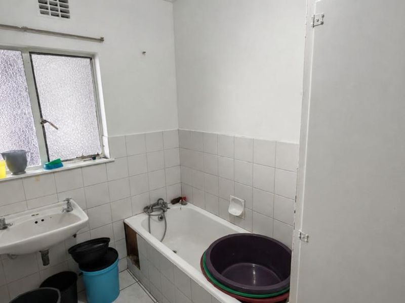 A functional bathroom featuring a bathtub, wall-mounted basin, and tiled finishes. The space requires cosmetic updates and repairs to the bathtub panel.