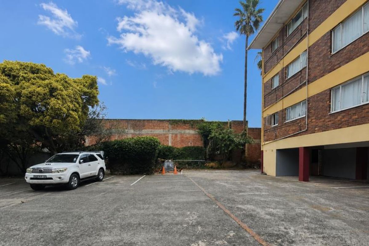 Exterior view of the apartment building showing a paved parking area, covered ground floor parking bays, and a perimeter boundary wall.
