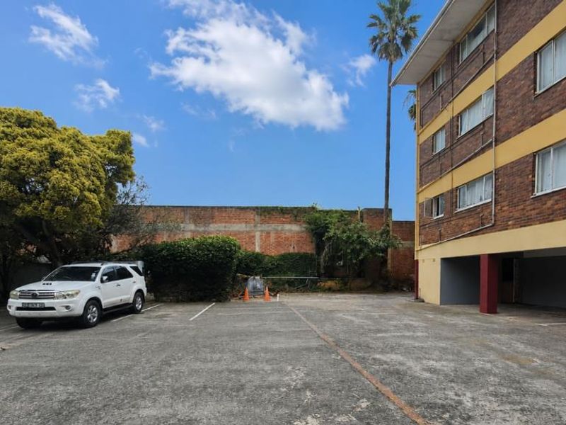 Exterior view of the apartment building showing a paved parking area, covered ground floor parking bays, and a perimeter boundary wall.