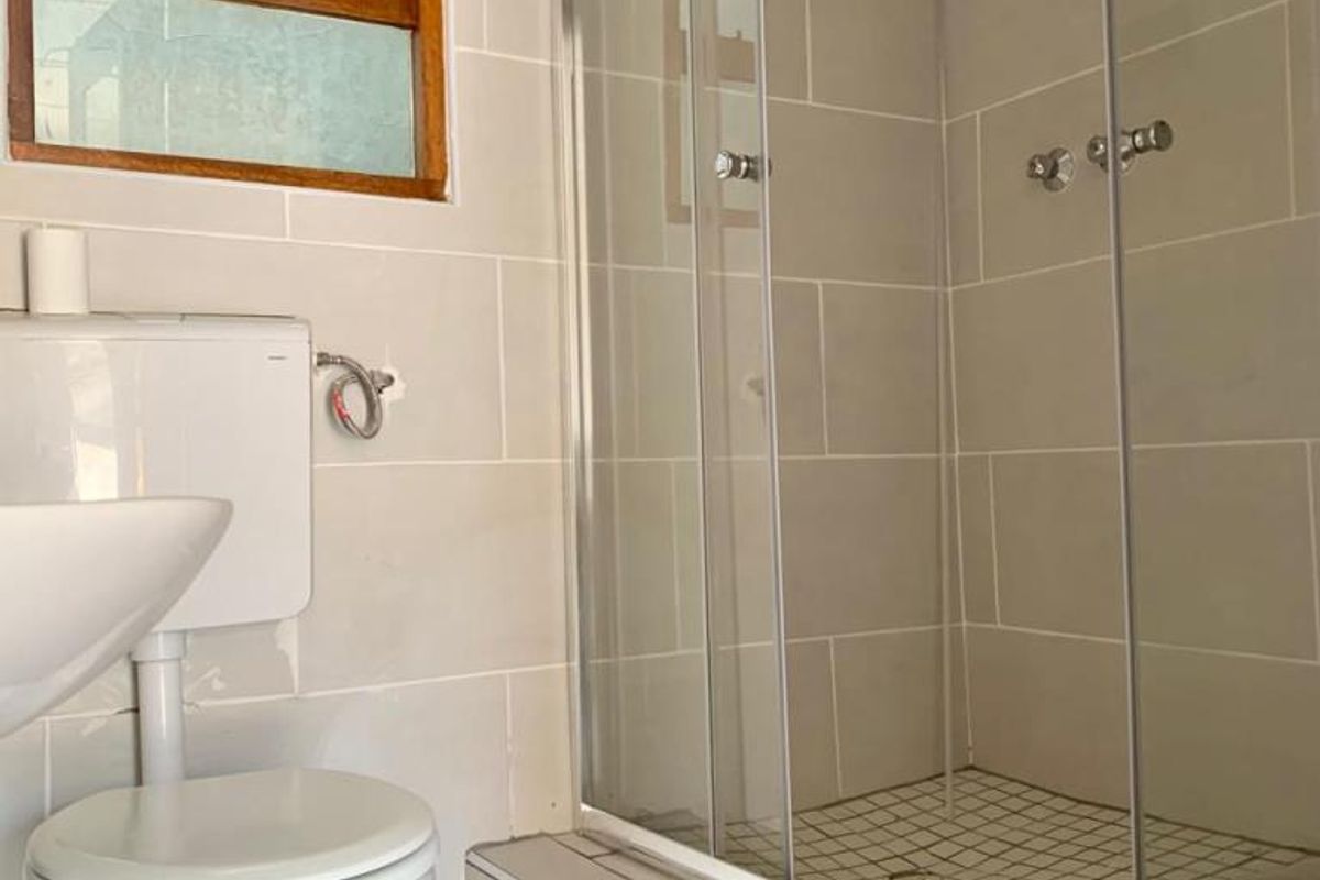 A functional bathroom featuring a glass-enclosed shower, white toilet, and washbasin with neutral wall tiling and a timber-framed window.