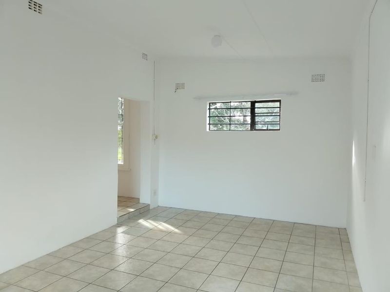 A bright, empty living area featuring neutral ceramic floor tiles, white walls, and a window fitted with security bars.