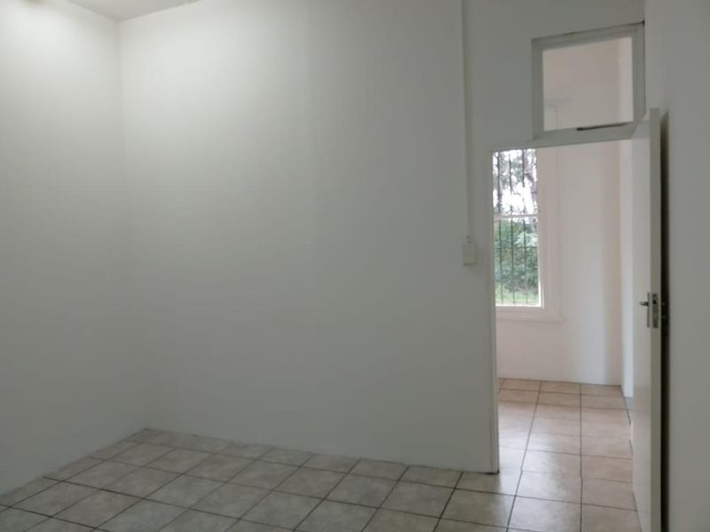 A clean, empty bedroom featuring neutral tiled flooring and a doorway leading to an adjacent space with security-barred windows.